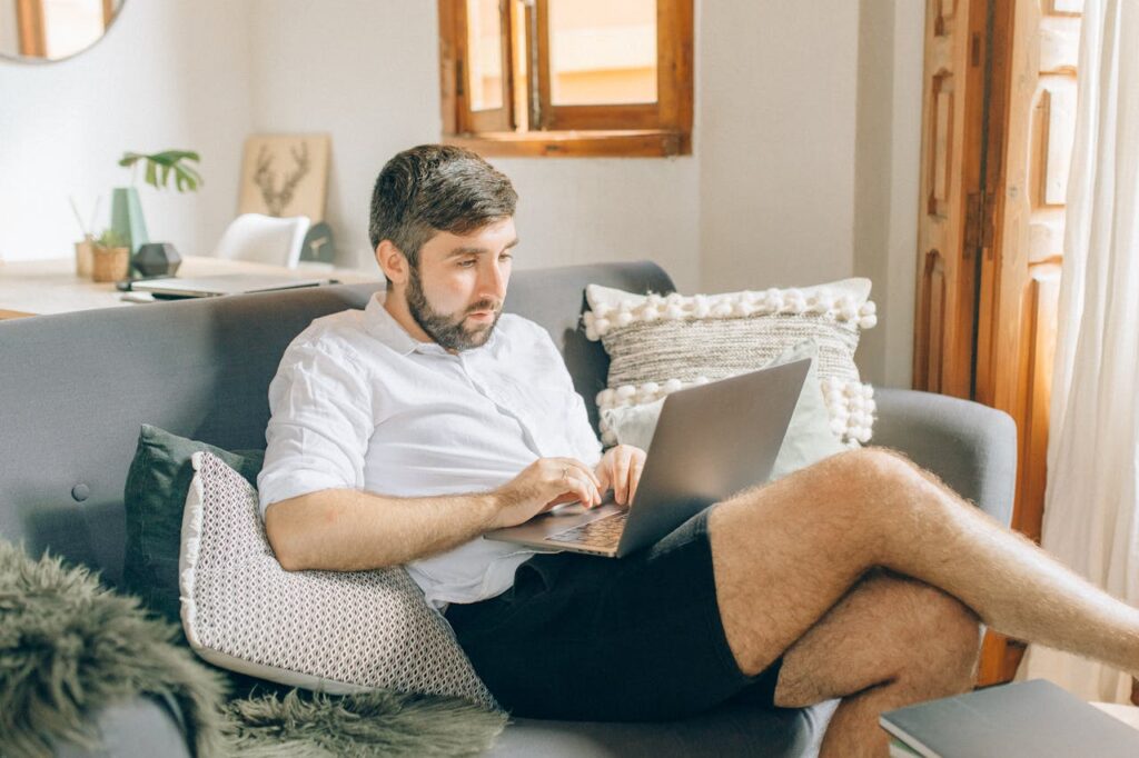 Adult man sitting on couch using laptop, working remotely from cozy home setting.